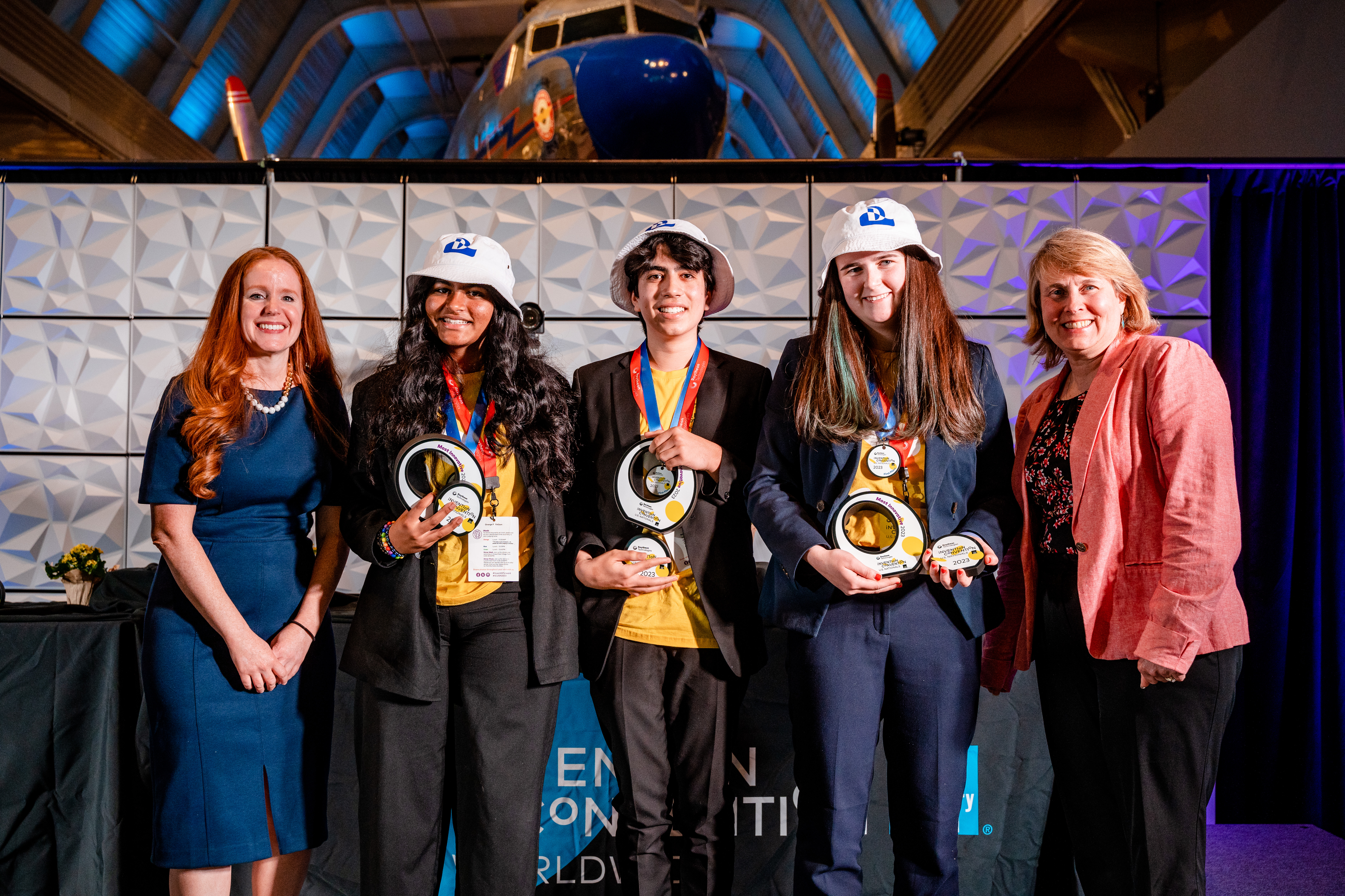 Three students in bucket hats accept circular awards on a stage