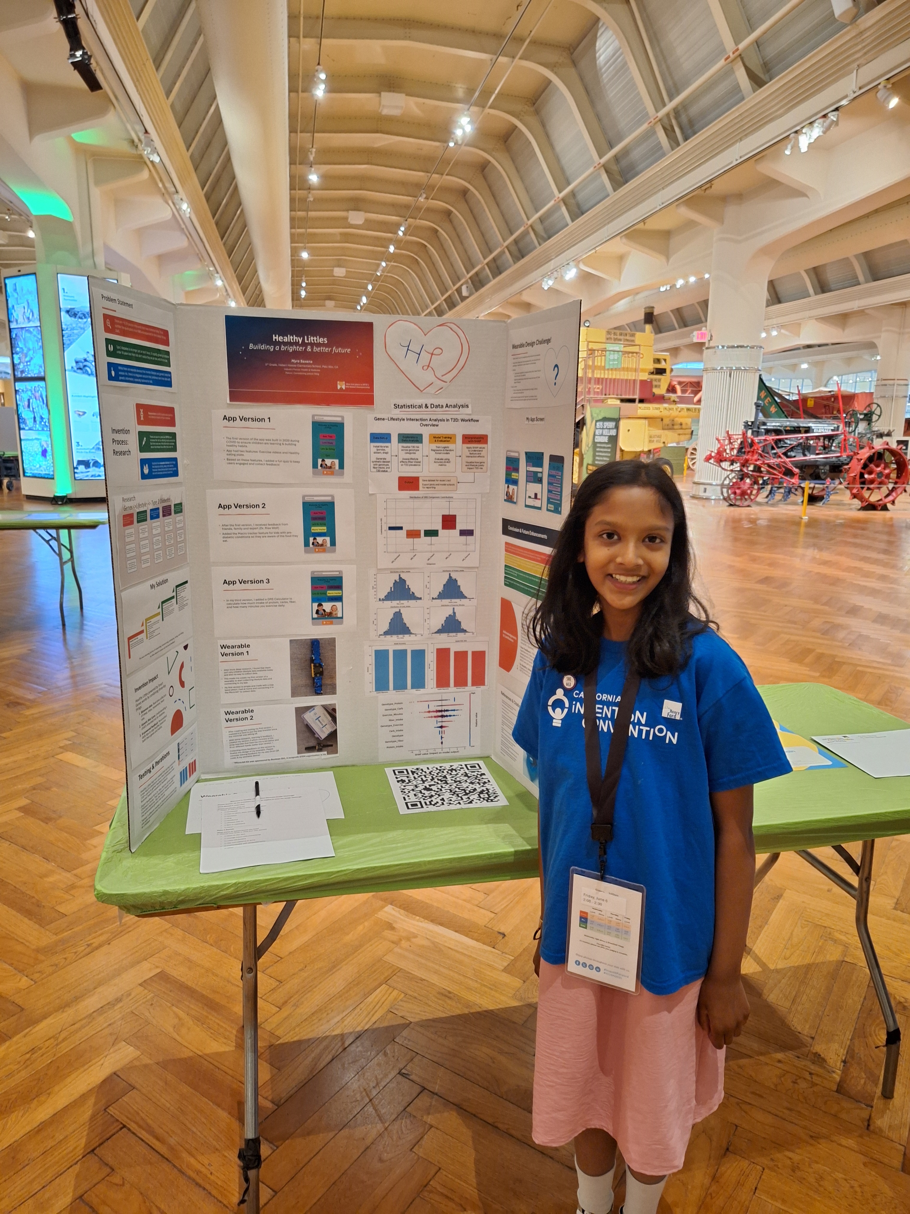A student in a blue t-shirt standing beside a tri-fold poster promoting her invention